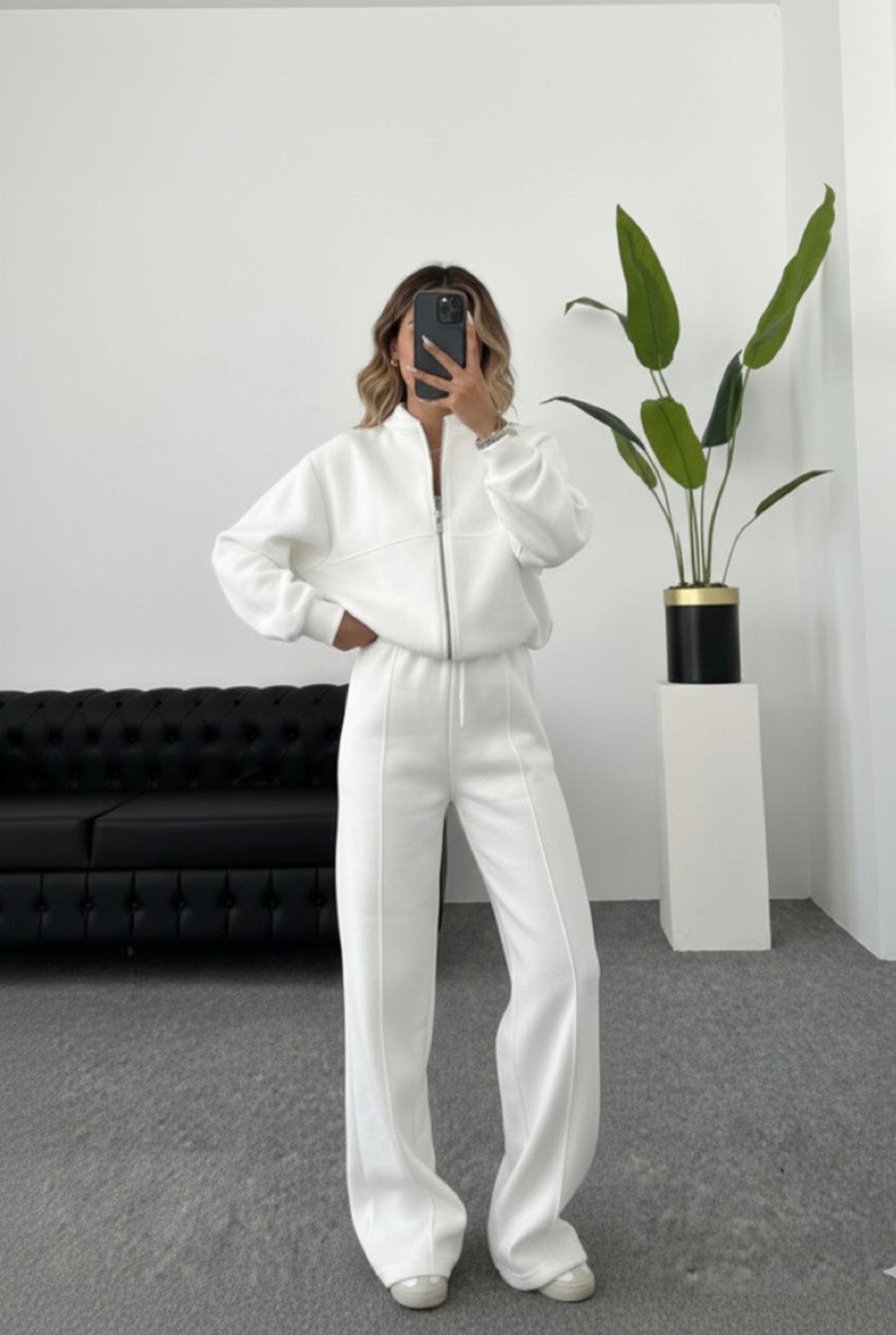 Person wearing a white tracksuit in a minimalistic room with a plant and black sofa.